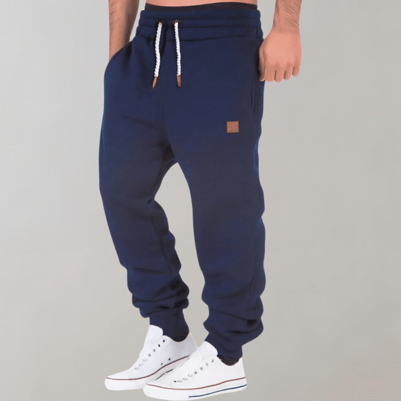 Donkerblauwe joggingbroek voor heren met witte trekkoorden en bruin logo-embleem, gecombineerd met witte sneakers. Comfortabele sportkleding.