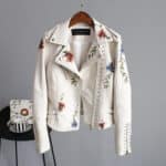 Beige leren jas met bloemenborduursels, zilveren studs en asymmetrische rits, hangend aan een houten hanger tegen een grijze achtergrond.