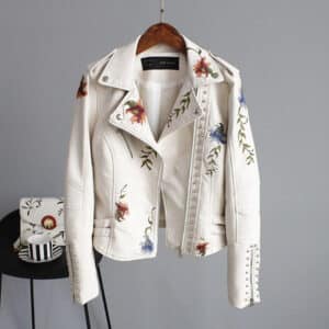 Beige leren jas met bloemenborduursels, zilveren studs en asymmetrische rits, hangend aan een houten hanger tegen een grijze achtergrond.