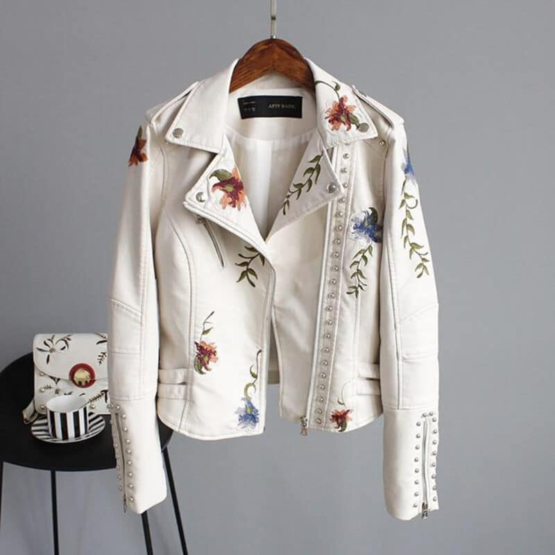 Beige leren jas met bloemenborduursels, zilveren studs en asymmetrische rits, hangend aan een houten hanger tegen een grijze achtergrond.