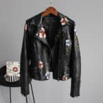 Zwarte leren jas met bloemenborduursels, studs en asymmetrische rits, hangend aan een houten hanger. Trendy damesmode, bikerstijl.