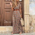 Vrouw in bohemian maxi-jurk met paisleyprint, bruine riem en strohoed, poseert voor houten deur. Zomerse mode, casual stijl, boho-chic outfit.