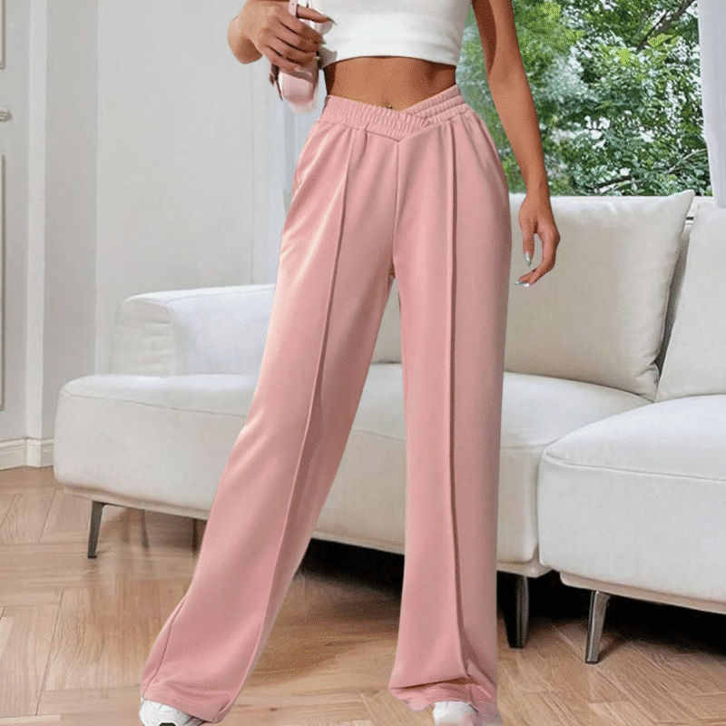 Vrouw in roze wijde broek en wit crop top, staand in moderne woonkamer. Trendy damesmode, comfortabele loungewear, stijlvolle outfit.