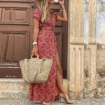 Vrouw in lange, donkerrode bohemian jurk met bloemenprint, draagt een strooien tas en sandalen, poseert voor houten deur. Zomerse mode, casual stijl.
