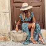 Vrouw in groene bohemian jurk met paisleyprint, zittend voor houten deur. Strohoed, rieten tas en transparante sandalen. Zomerse mode, boho stijl.