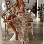 Vrouw in lange feestelijke jurk met bladprint, zittend aan houten tafel in moderne eetkamer. Trendy damesmode, casual chique stijl.