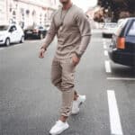 Man in beige trainingspak loopt op straat, draagt witte sneakers en horloge. Stijlvolle herenkleding, casual mode, stedelijke achtergrond.