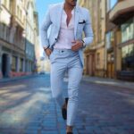 Stijlvolle man in lichtblauw slim fit pak, witte overhemd en zwarte loafers, wandelend in stedelijke straat. Modieuze herenmode, elegante uitstraling.