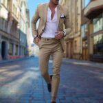 Stijlvolle man in beige slim fit pak, witte overhemd en zwarte loafers, lopend op geplaveide straat. Modieuze herenkleding, stedelijke achtergrond.