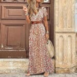 Vrouw in lange, bloemenprint zomerjurk met riem, rieten tas en hoed voor houten deur. Perfecte boho-chic outfit voor warme dagen.