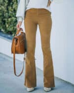 Vrouw draagt bruine flared broek met witte trui, houdt bruine leren tas vast. Modieuze herfstoutfit, casual stijl, trendy damesmode.