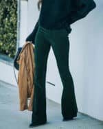 Vrouw draagt donkergroene flared broek met zwarte trui, houdt bruine jas vast. Modieuze herfstoutfit, trendy dameskleding, straatstijl.